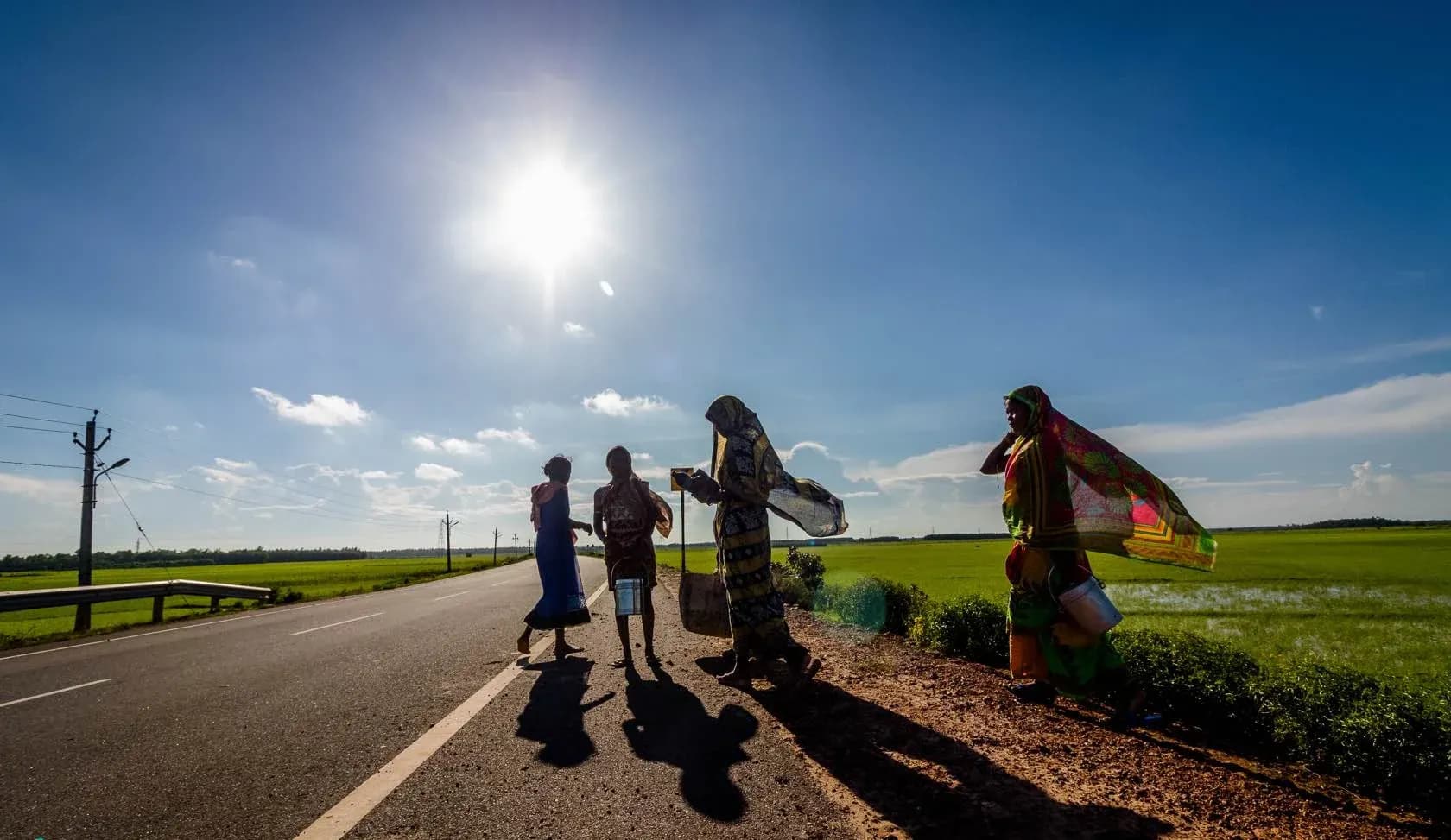 Local women walking along a sunlit road through green fields, silhouetted against the sun